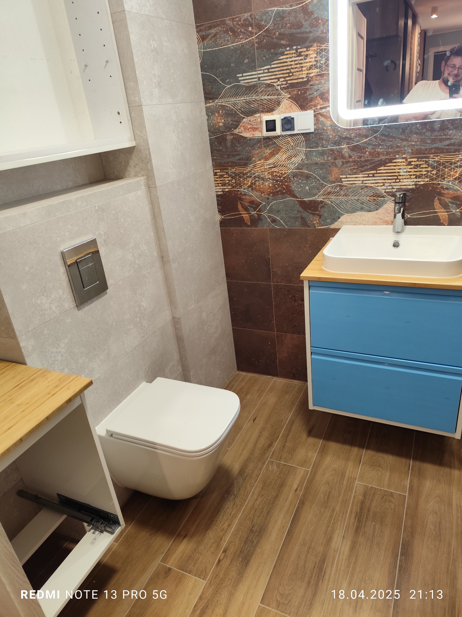 Nowoczesna łazienka po remoncie: wisząca toaleta, niebieska szafka z umywalką, lustro z podświetleniem LED, brązowe płytki z motywem liści i szare płytki.