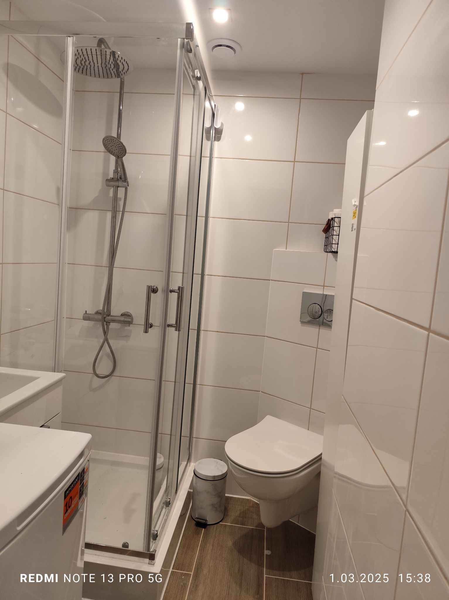 Jasna łazienka po renowacji: kabina prysznicowa ze szklanymi drzwiami, toaleta i białe płytki na ścianach. Widoczne detale wykończenia i armatura.