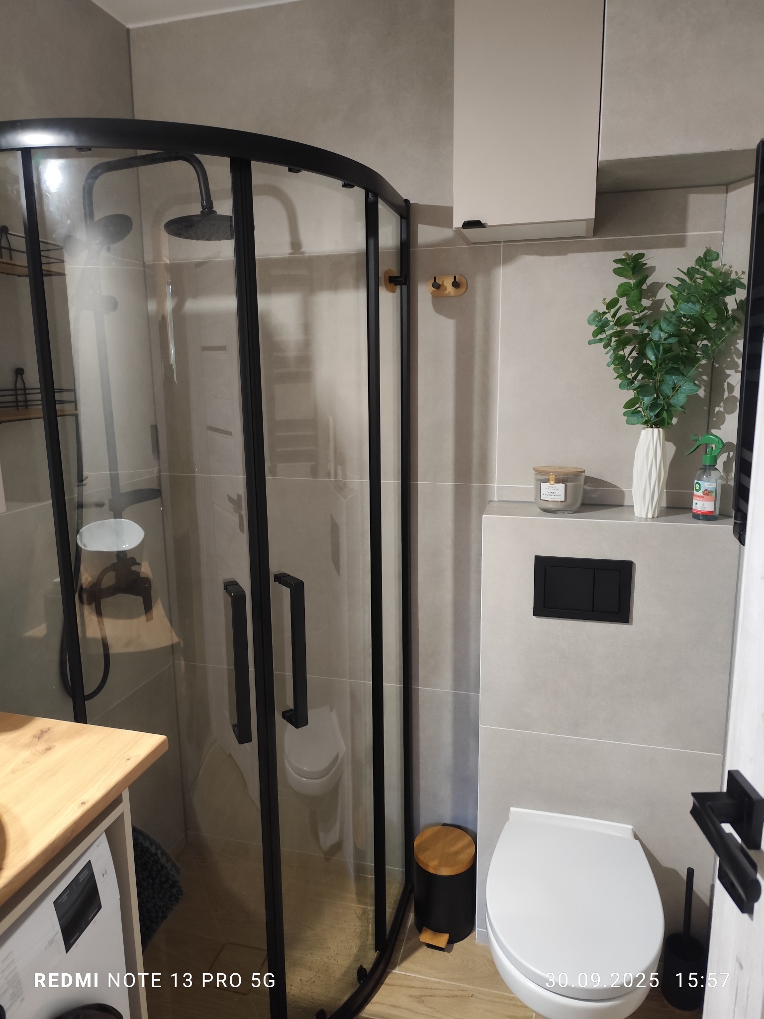 Nowoczesna łazienka w Pruszczu Gdańskim z kabiną prysznicową typu walk-in z czarnymi profilami, jasnymi płytkami i drewnianym blatem. Minimalistyczny design.