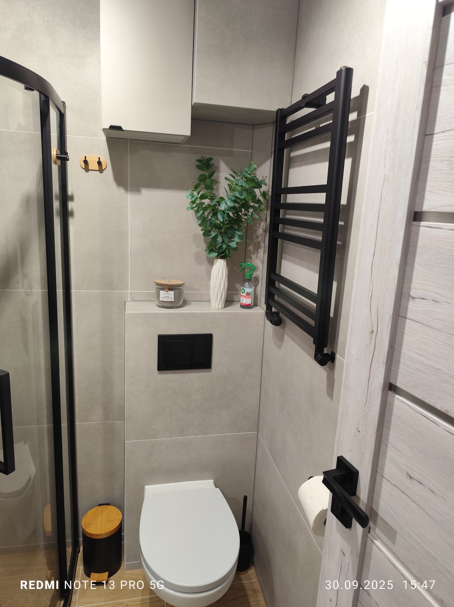 Nowoczesna łazienka w szarościach z czarnymi akcentami: grzejnik drabinkowy, kabina prysznicowa, toaleta podwieszana i dekoracje. Minimalistyczny design z roślinami.