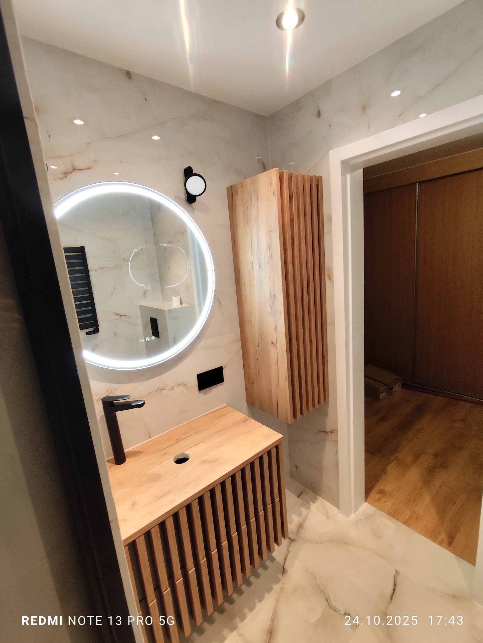 Nowoczesna łazienka po remoncie: drewniana szafka z umywalką, okrągłe lustro z podświetleniem LED i wisząca szafka z lamelami na tle marmurowych płytek.