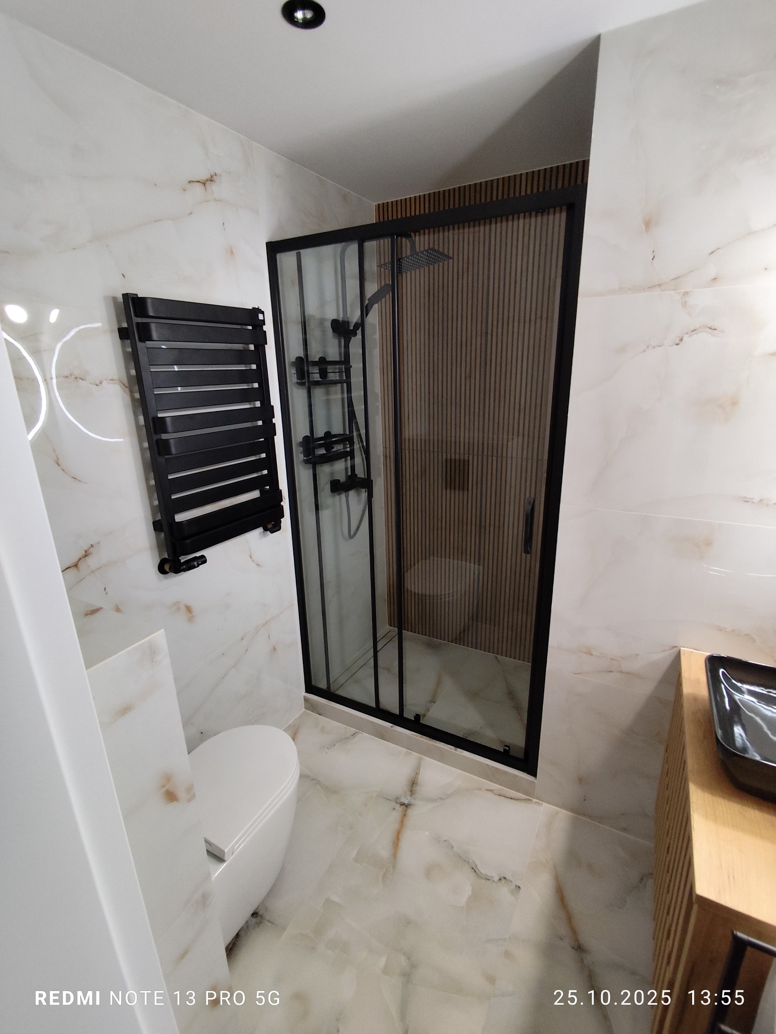 Nowoczesna łazienka z kabiną prysznicową w czarnej ramie, marmurowe płytki na ścianach i podłodze, grzejnik drabinkowy i toaleta. Minimalistyczny design.