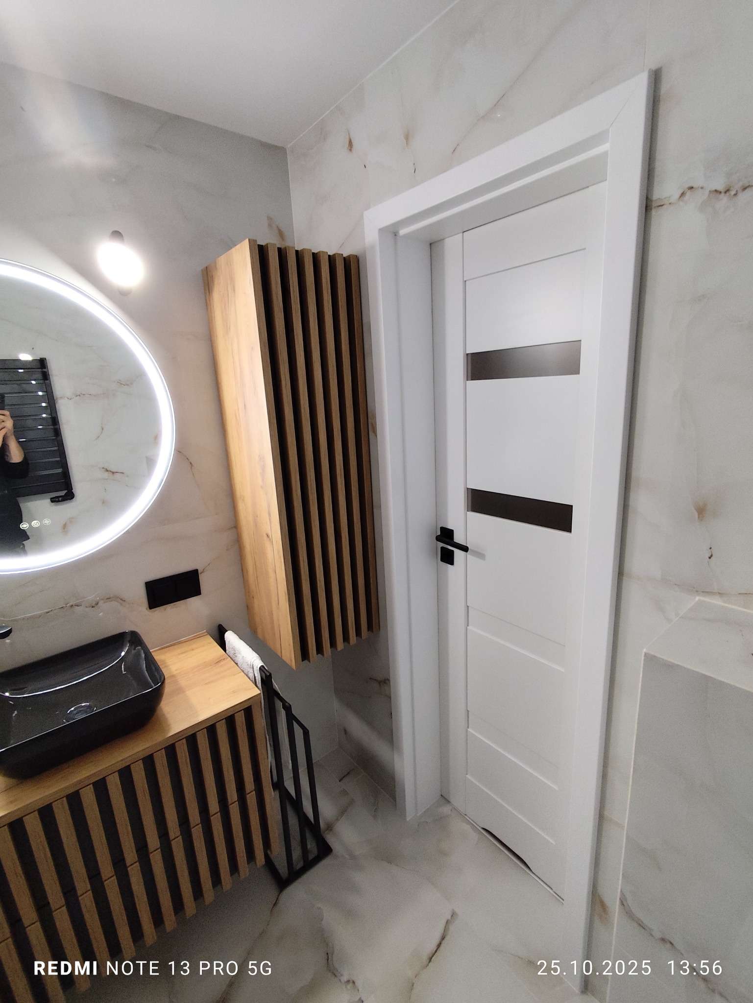 Nowoczesna łazienka z umywalką nablatową, szafką z drewnianymi lamelami i okrągłym lustrem z podświetleniem LED. Marmurowe płytki na ścianach i podłodze. Białe drzwi z czarną klamką.
