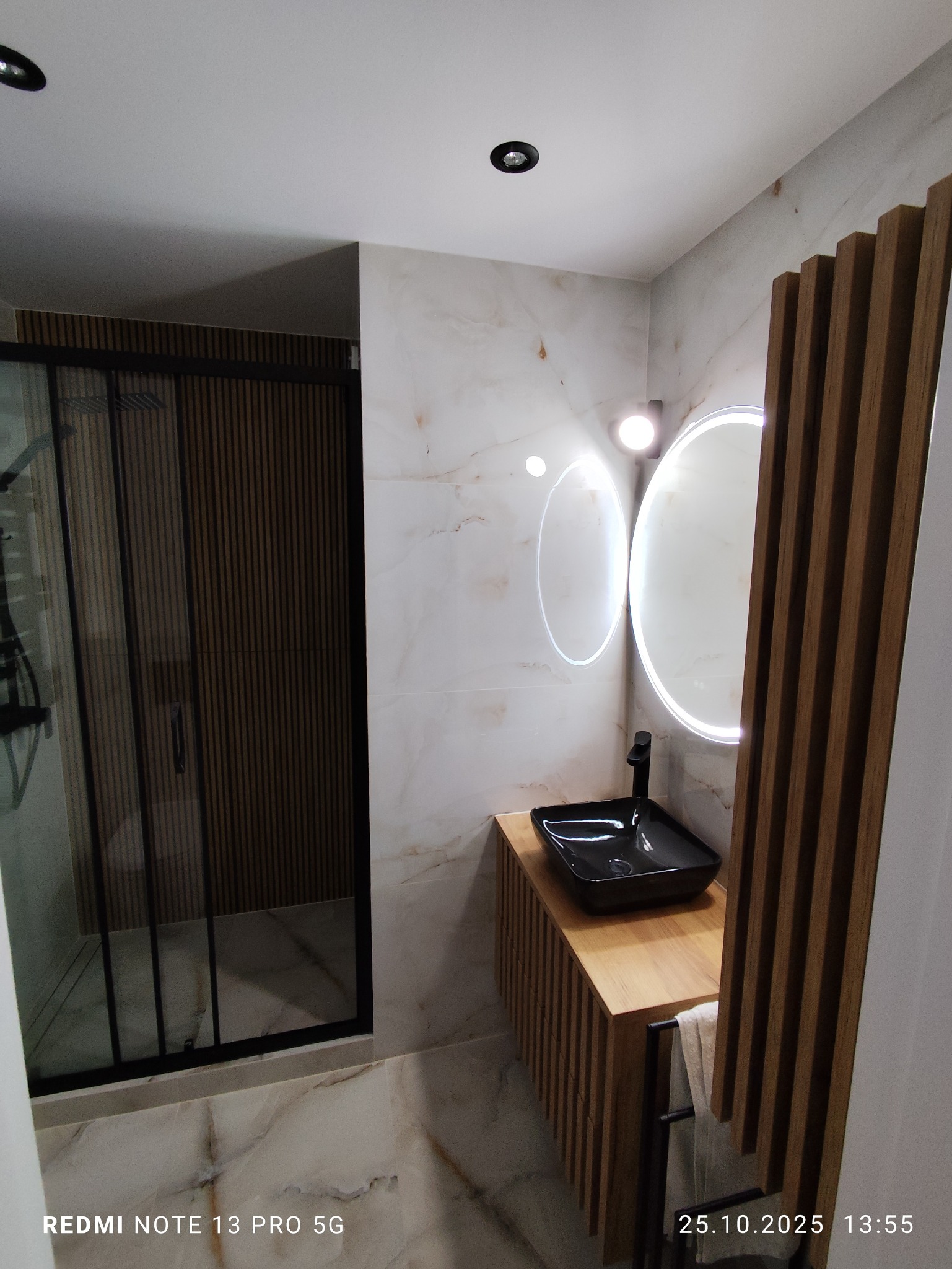 Nowoczesna łazienka z kabiną prysznicową typu walk-in, umywalką nablatową i podświetlanym lustrem. Marmurowe płytki i drewniane akcenty tworzą elegancki wystrój.