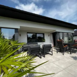 Biała elewacja domu z czarnymi oknami i dachem, widok z tarasu z szarymi płytkami i meblami ogrodowymi, zieleń roślin na pierwszym planie.