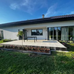 Nowoczesny dom jednorodzinny z białymi ścianami, dużymi oknami i tarasem wyłożonym jasnymi płytami, otoczony zielonym trawnikiem i rabatami z czerwonymi kwiatami w słoneczny dzień.