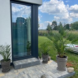 Agencja doradztwa energetycznego Dawid Lipko - Nowoczesny dom z szarą elewacją, dużym oknem z ciemną ramą i wybrukowanym tarasem, na którym stoją donice z palmami i dekoracyjne kamienie.