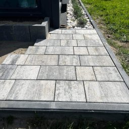 Agencja doradztwa energetycznego Dawid Lipko - Ułożona z szarych kostek brukowych ścieżka prowadząca do drzwi budynku, obok rabata z białymi kamieniami i zielonymi roślinami, widok z poziomu gruntu.