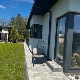 Agencja doradztwa energetycznego Dawid Lipko - Nowoczesny dom z elewacją w jasnym odcieniu, widoczne okna z ciemnymi ramami, chodnik z kostki brukowej, trawnik i krzesło ogrodowe