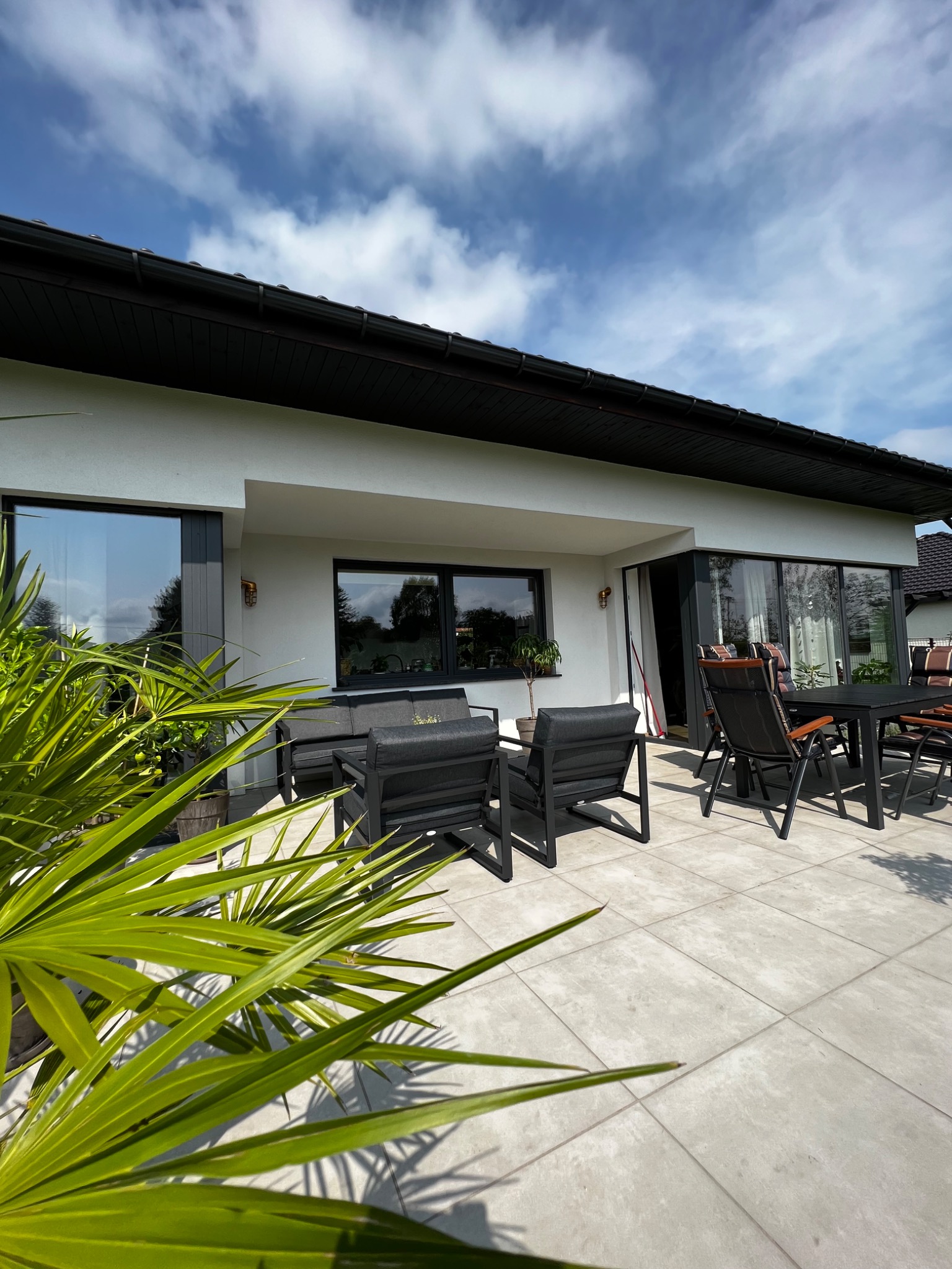Biała elewacja domu z czarnymi oknami i dachem, widok z tarasu z szarymi płytkami i meblami ogrodowymi, zieleń roślin na pierwszym planie.