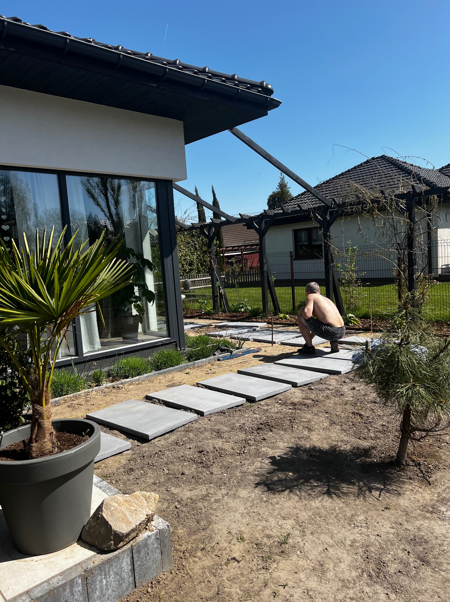 Mężczyzna w krótkich spodenkach układa betonowe płyty chodnikowe w ogrodzie przed nowoczesnym domem z dużymi oknami, widoczny palmowiec w donicy i pergola.
