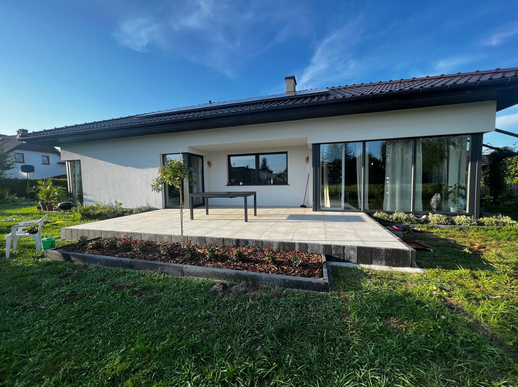 Nowoczesny dom jednorodzinny z białymi ścianami, dużymi oknami i tarasem wyłożonym jasnymi płytami, otoczony zielonym trawnikiem i rabatami z czerwonymi kwiatami w słoneczny dzień.