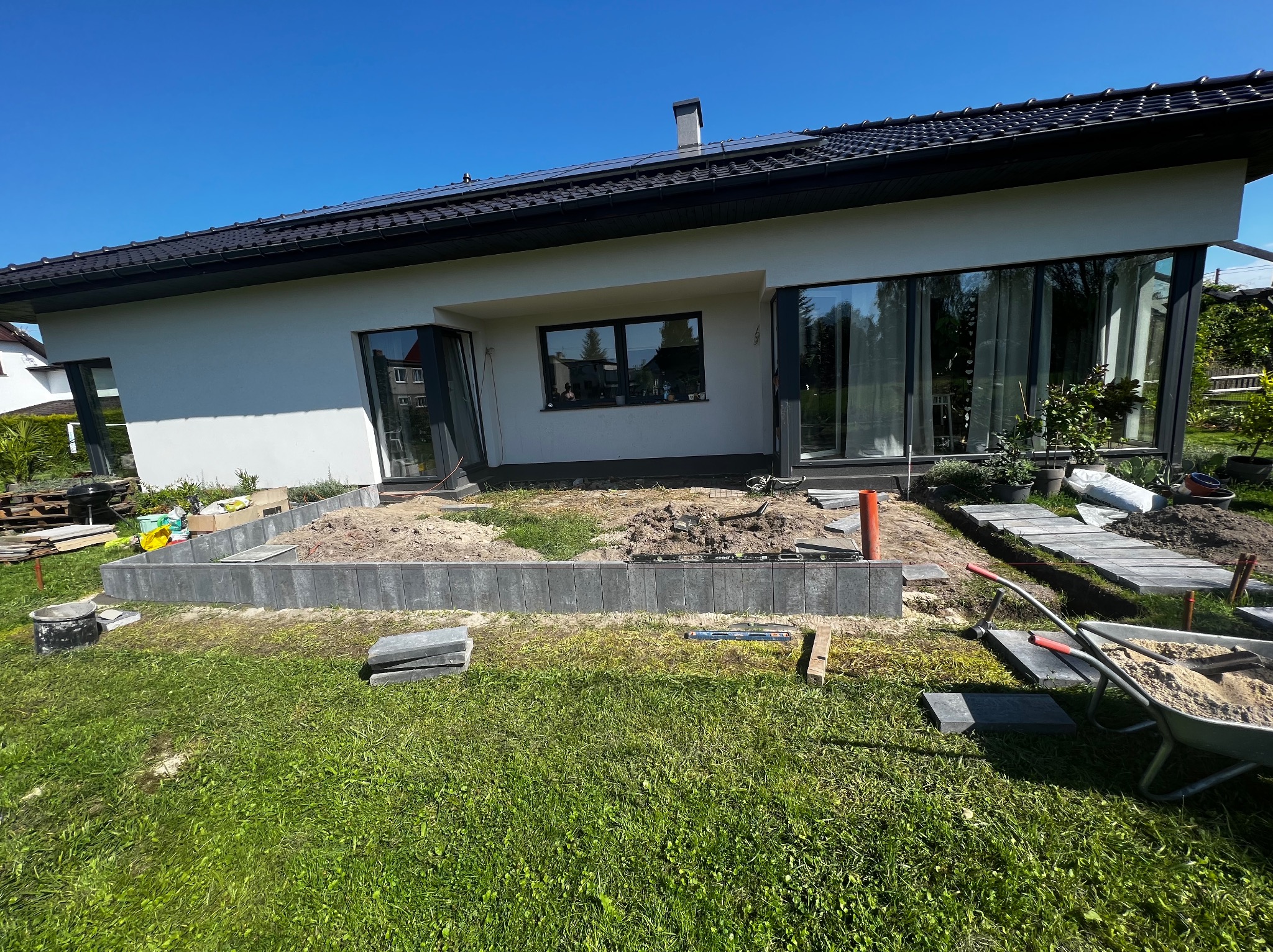 Nowoczesny dom jednorodzinny z białymi ścianami i czarną dachówką w trakcie budowy rabaty kwiatowej z szarych bloczków betonowych, widoczny wózek z piaskiem i narzędzia.