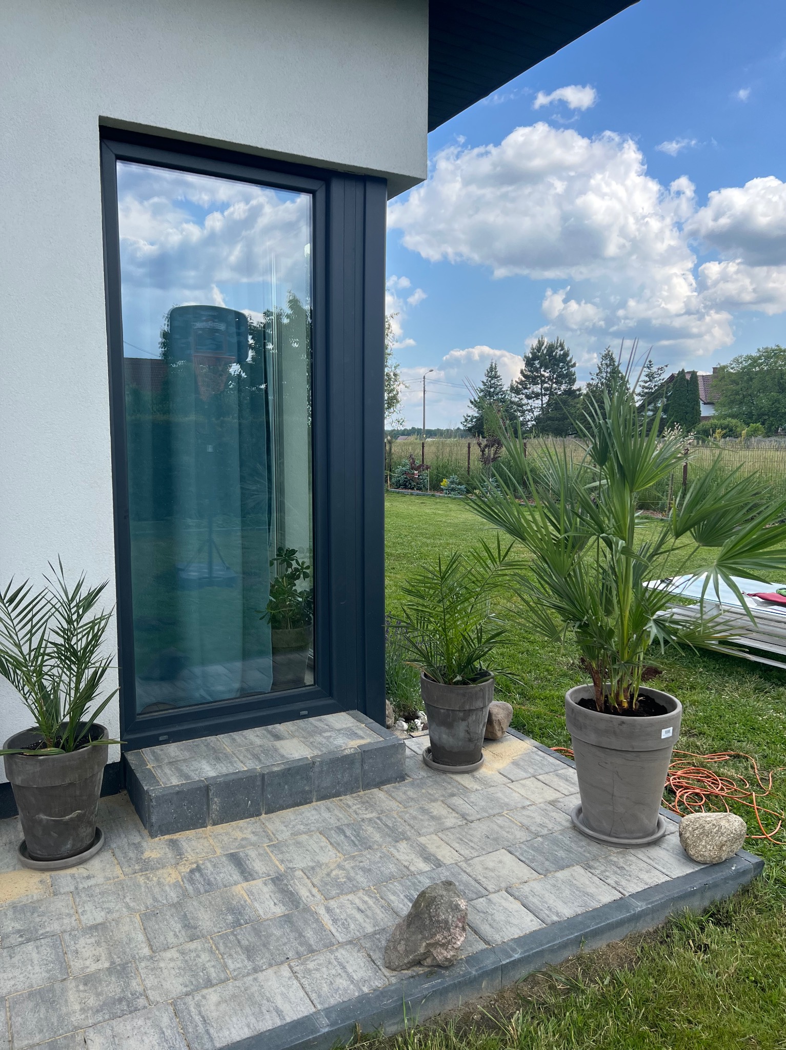 Nowoczesny dom z szarą elewacją, dużym oknem z ciemną ramą i wybrukowanym tarasem, na którym stoją donice z palmami i dekoracyjne kamienie.