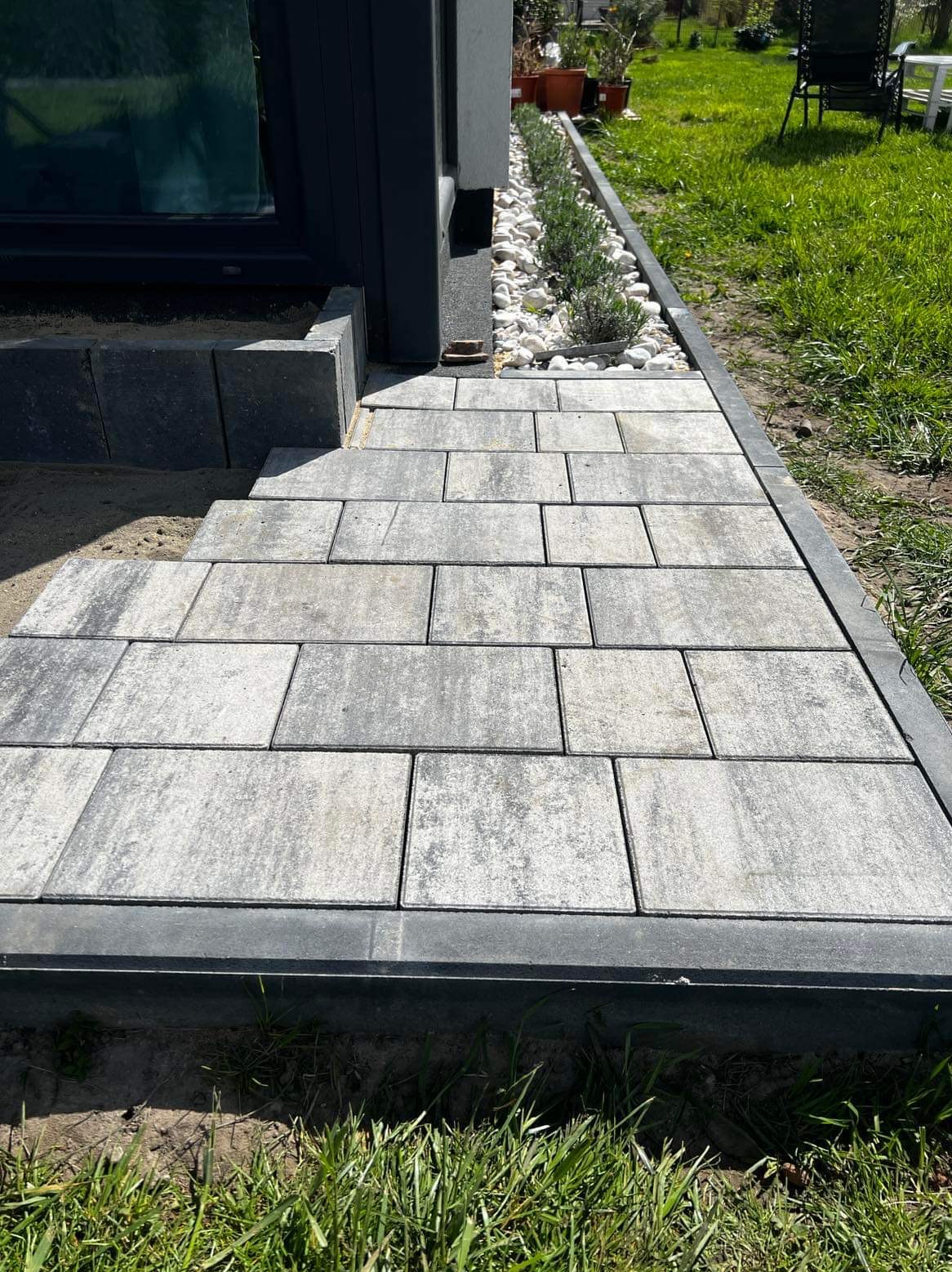 Ułożona z szarych kostek brukowych ścieżka prowadząca do drzwi budynku, obok rabata z białymi kamieniami i zielonymi roślinami, widok z poziomu gruntu.