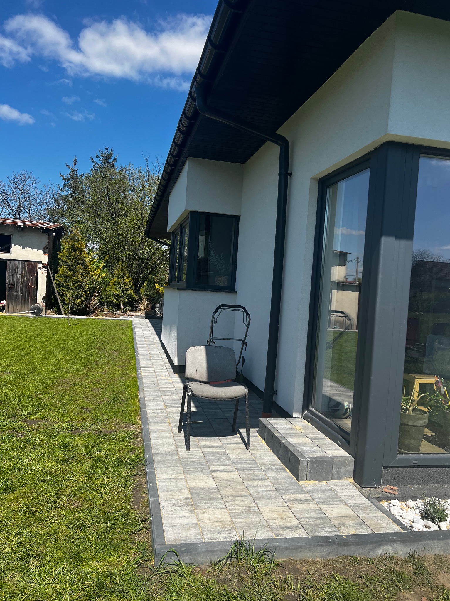 Nowoczesny dom z elewacją w jasnym odcieniu, widoczne okna z ciemnymi ramami, chodnik z kostki brukowej, trawnik i krzesło ogrodowe