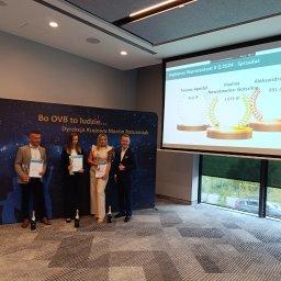 FinanSecure Paulina Nowakowska-Gotszling - Wręczenie nagród dla najlepszych reprezentantów firmy OVB w II kwartale 2024, na tle baneru firmowego i wyświetlanego slajdu z wynikami, z szampanem na podłodze.