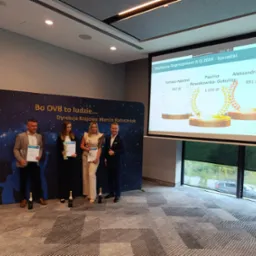 Wręczenie nagród dla najlepszych reprezentantów firmy OVB w II kwartale 2024, na tle baneru firmowego i wyświetlanego slajdu z wynikami, z szampanem na podłodze.