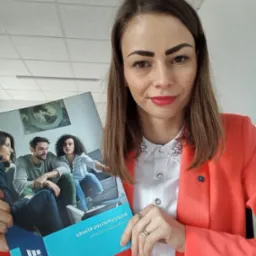 Kobieta w koralowej marynarce prezentuje broszurę z wizerunkiem rodziny na kanapie i tekstem 'Analiza potrzeb klienta + CHECK-UP' odwróconym do góry nogami.