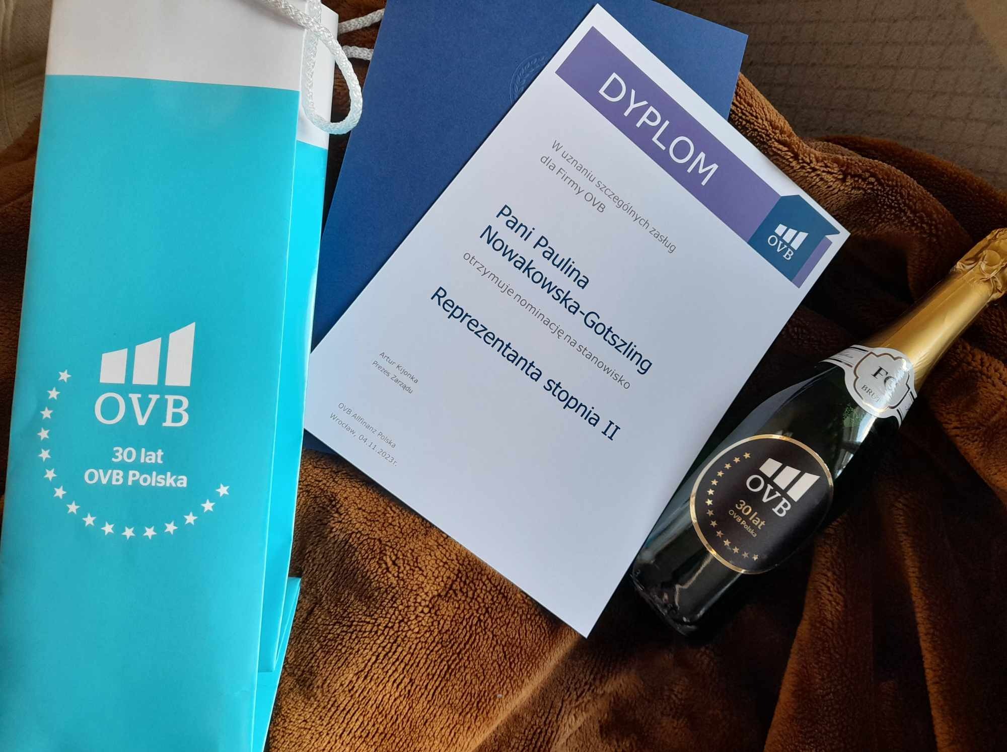 Dyplom dla Pani Pauliny Nowakowskiej-Gotszling za zasługi dla firmy OVB, prezentowany obok torby z logo firmy i butelki szampana z okolicznościową etykietą, całość ułożona na brązowym tle.