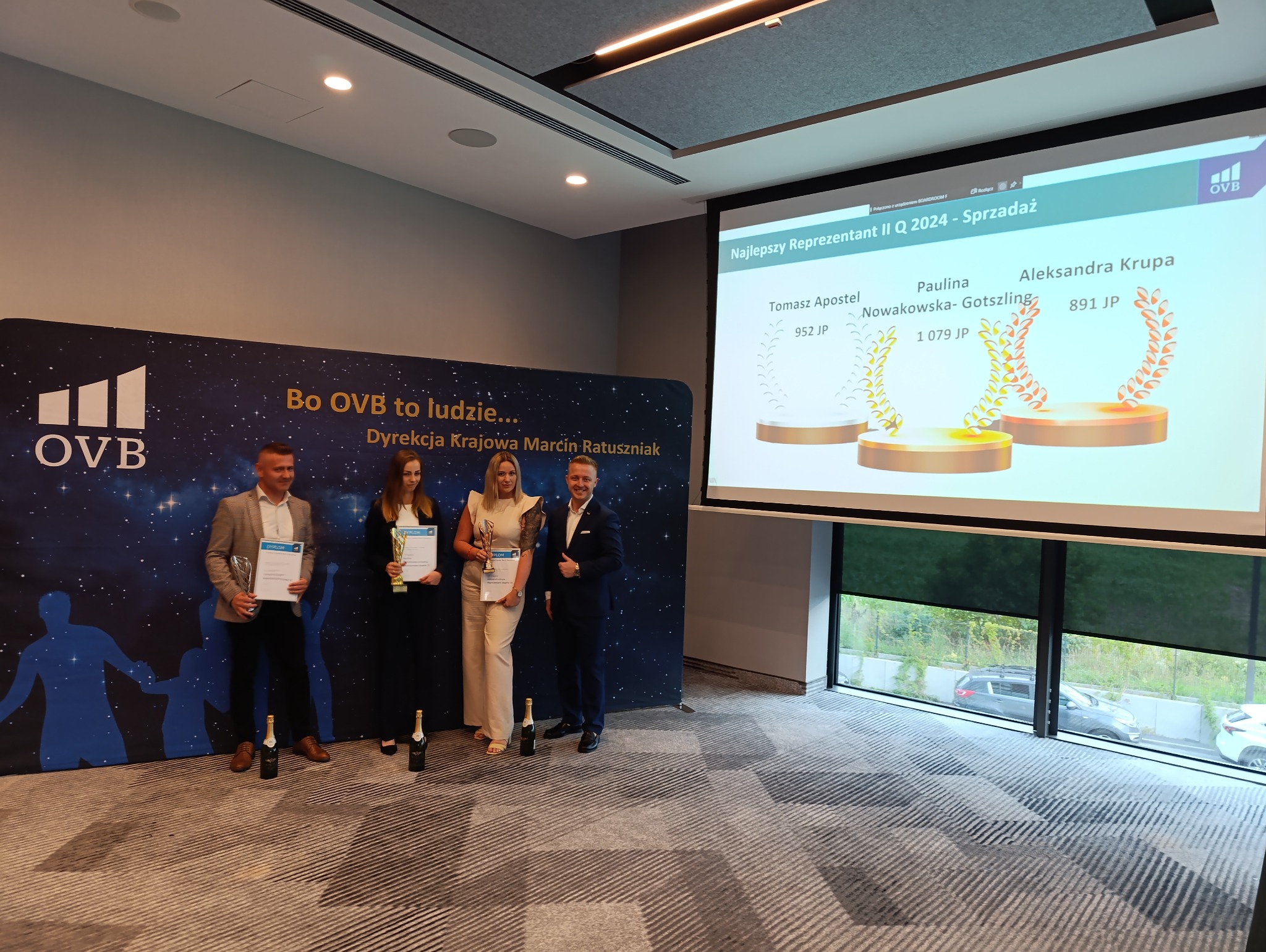 Wręczenie nagród dla najlepszych reprezentantów firmy OVB w II kwartale 2024, na tle baneru firmowego i wyświetlanego slajdu z wynikami, z szampanem na podłodze.