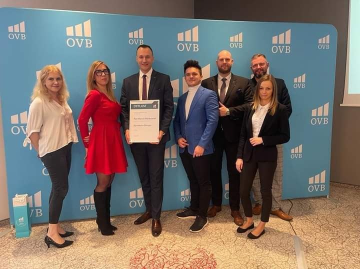 Zespół z Rawicza świętuje sukces, mężczyzna w garniturze trzyma dyplom na tle ściany z logo OVB, obok stoją współpracownicy.