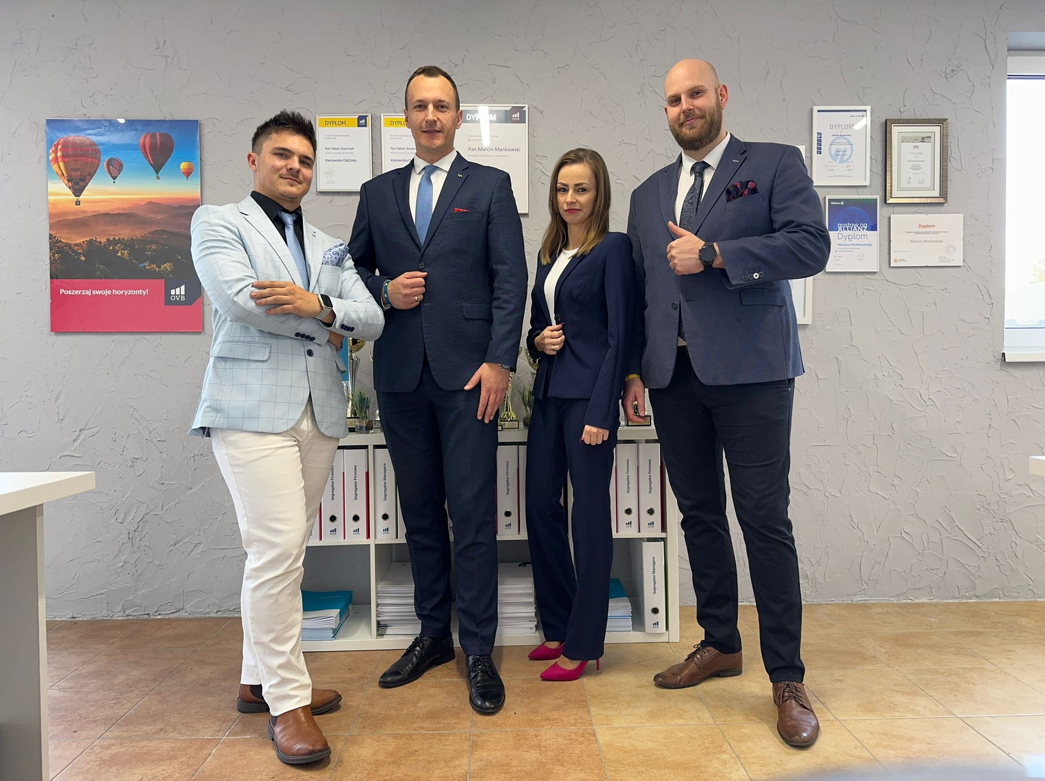 Grupa czterech osób w garniturach pozuje w biurze, na ścianie dyplomy i plakat z balonami, na półce segregatory z napisami 'Segregator Firmowy' i 'Segregator Managera'.