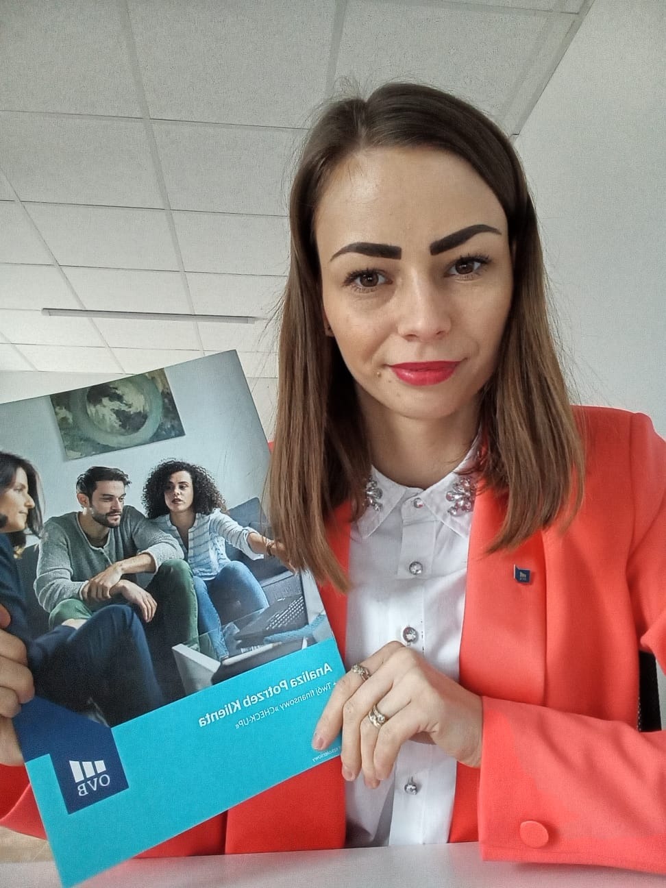 Kobieta w koralowej marynarce prezentuje broszurę z wizerunkiem rodziny na kanapie i tekstem 'Analiza potrzeb klienta + CHECK-UP' odwróconym do góry nogami.