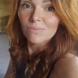 Portret kobiety z rudymi, kręconymi włosami i delikatnym makijażem, noszącej naszyjnik z zawieszką w kształcie liścia.