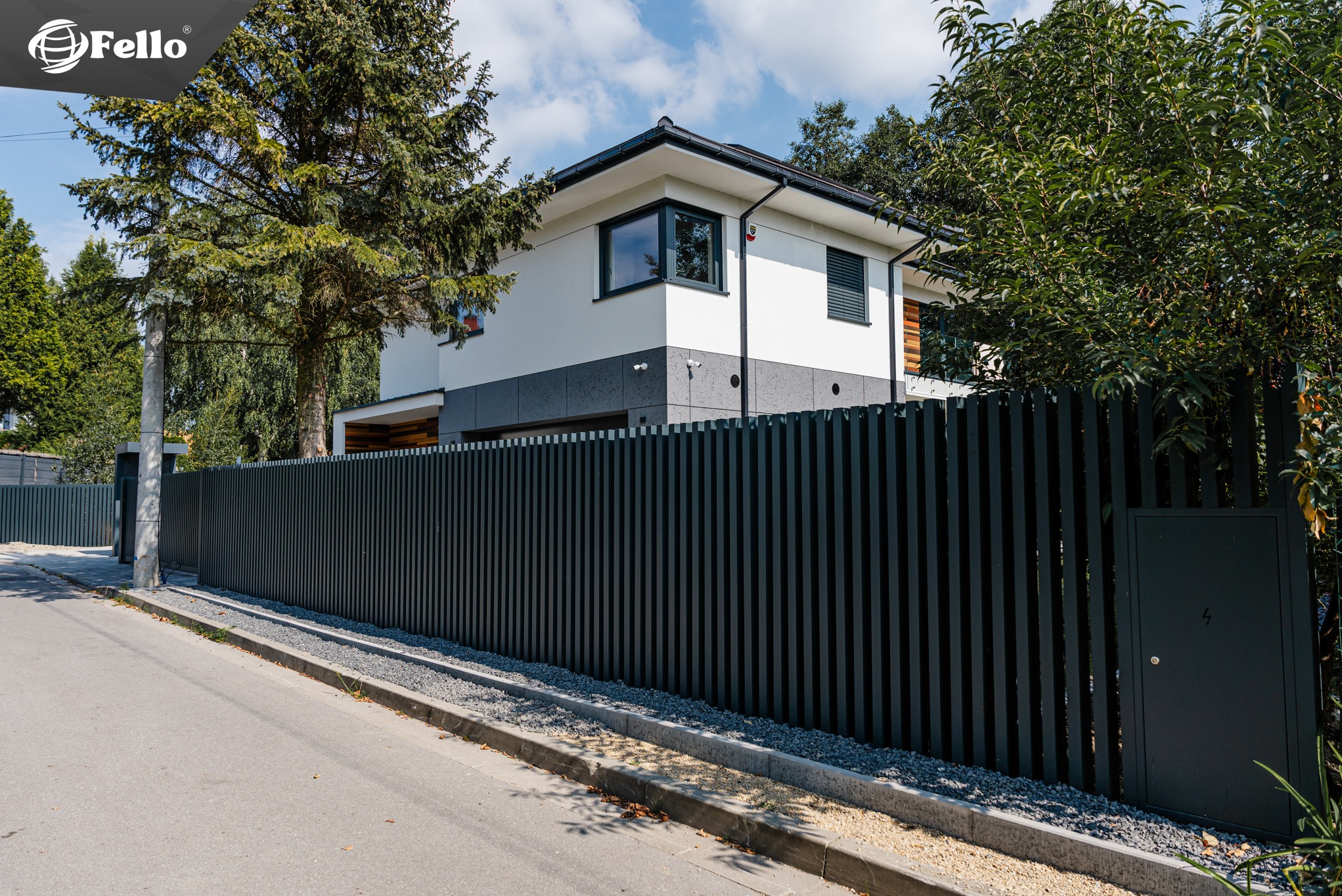 Nowoczesne ogrodzenie metalowe typu vertical w kolorze grafitowym, z pionowymi panelami, na tle dwukondygnacyjnego domu z białymi i szarymi elementami elewacji.