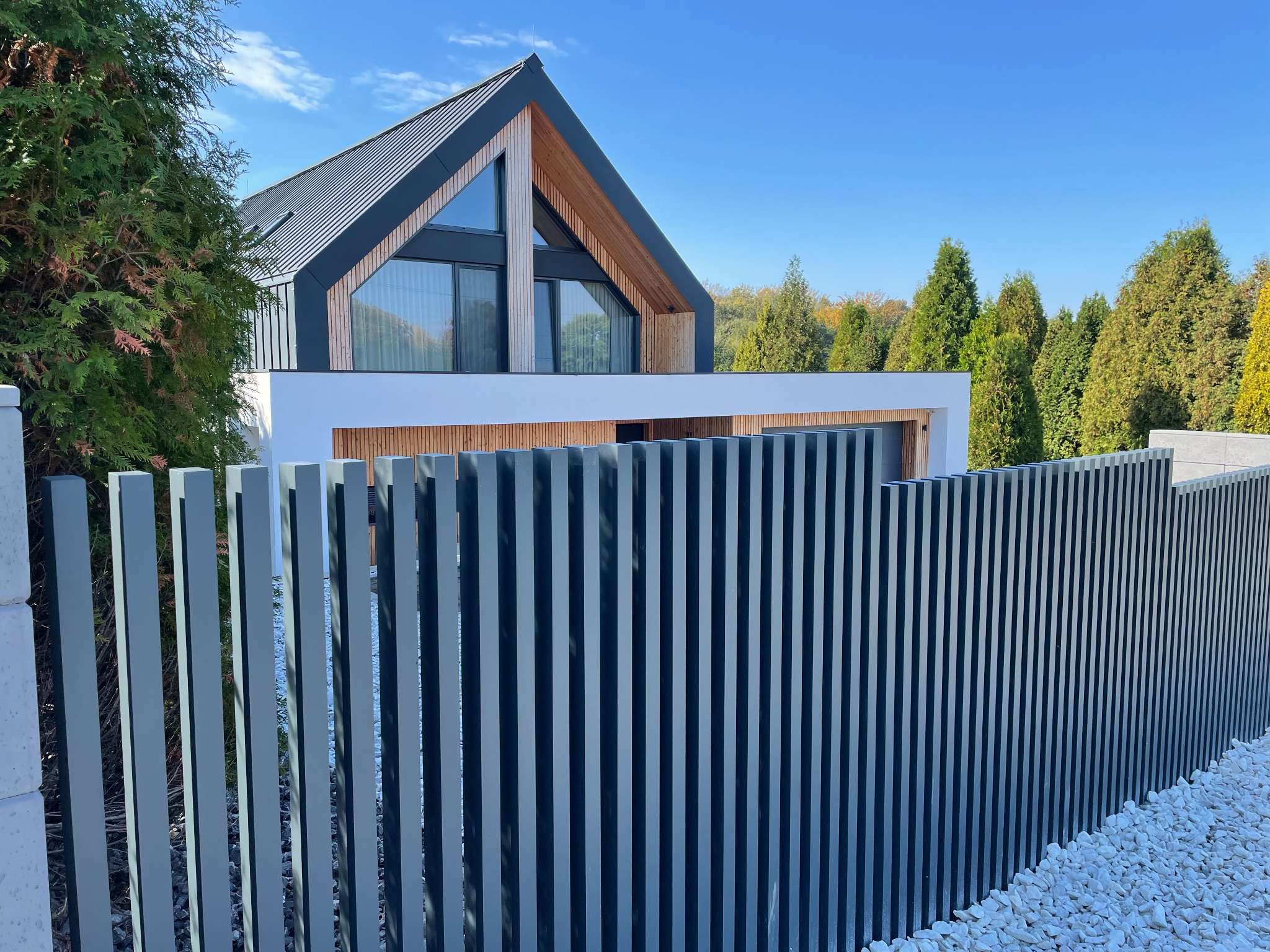 Nowoczesne ogrodzenie metalowe typu vertical w kolorze grafitowym, z widokiem na dom i ozdobny żwir. Słoneczny dzień.