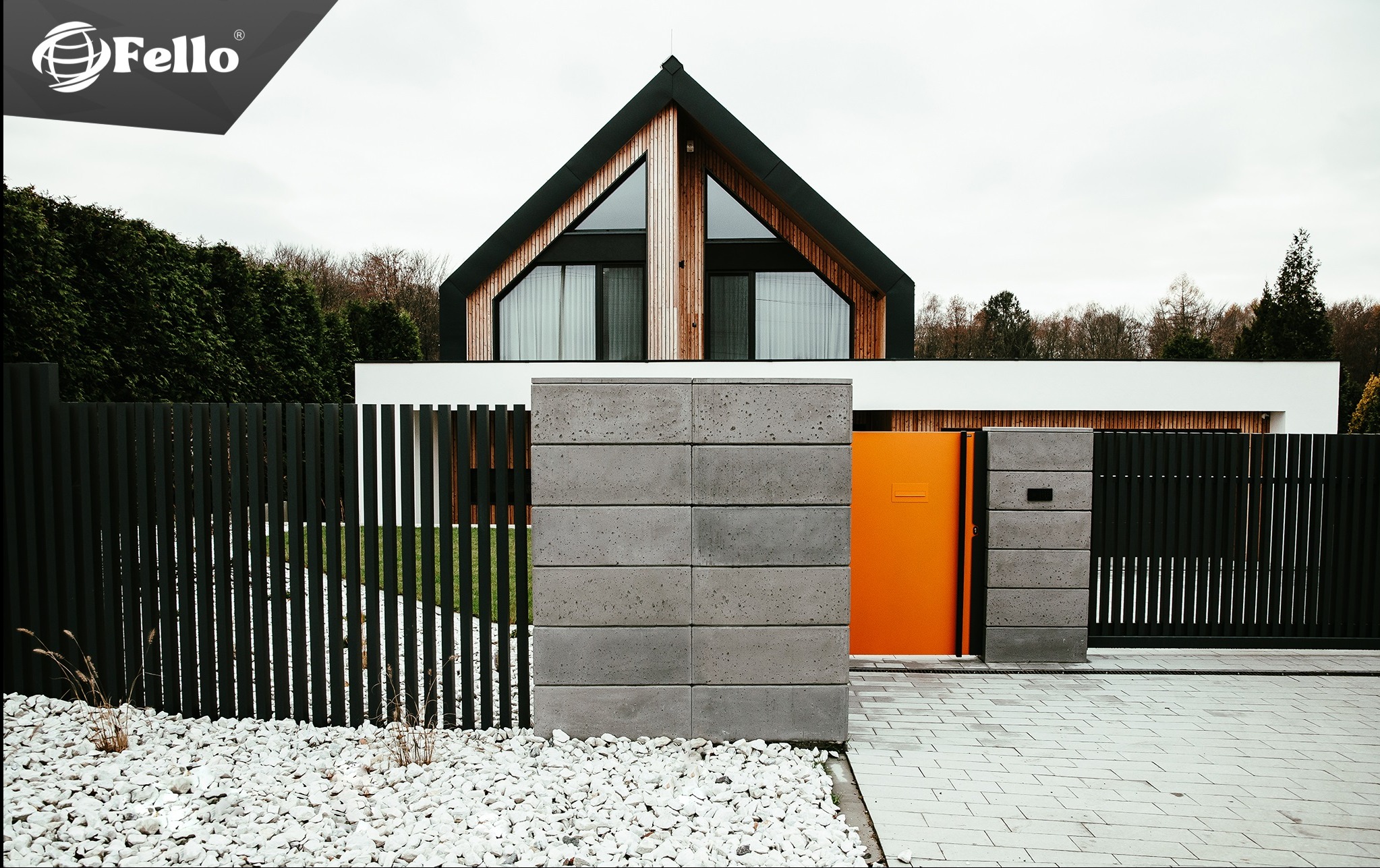 Nowoczesne ogrodzenie panelowe w kolorze grafitowym z pomarańczową bramą na tle domu o architekturze stodoły, otoczone białymi kamieniami.