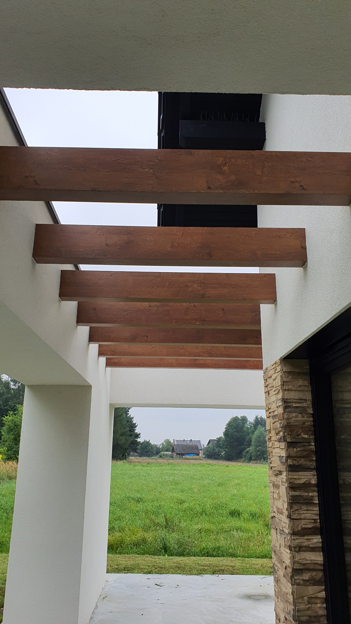 Drewnopodobne belki dekoracyjne pod sufitem nowoczesnej pergoli, widok na zielony krajobraz. Minimalistyczny design, neutralna kolorystyka, kamienna kolumna.