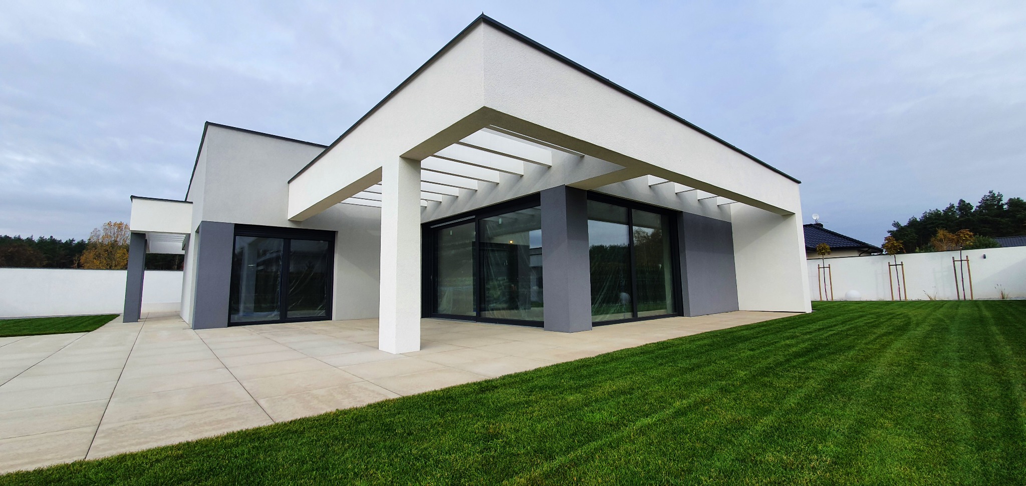 Nowoczesna pergola metalowa z belkami dekoracyjnymi przy domu z dużymi oknami przesuwnymi, widok na trawnik i ogrodzenie. Minimalistyczny design.