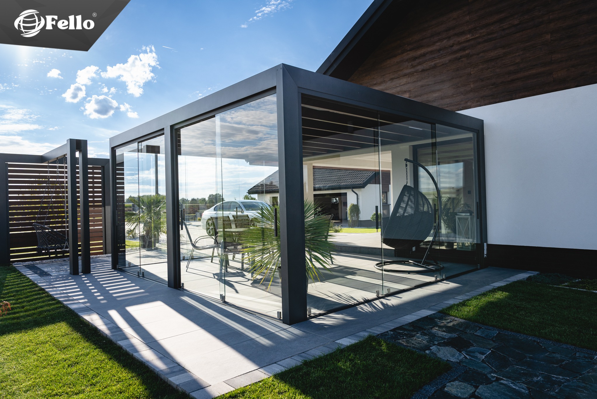 Nowoczesna, przeszklona pergola z ciemną ramą aluminiową, wyposażona w wiszące krzesło i rośliny, na tle zadbanego ogrodu z domem jednorodzinnym w tle.