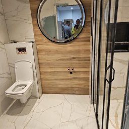 Damiano - Nowoczesna łazienka w trakcie wykańczania: białe marmurowe płytki podłogowe i ścienne, drewnopodobna okładzina ściany z okrągłym lustrem, podwieszana toaleta i czarna kabina prysznicowa z widocznym...