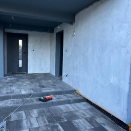 Damiano - Surowy stan werandy z szarymi płytami chodnikowymi, ściany w kolorze szarym, widoczne narzędzia glazurnicze i kable, w tle drzwi wejściowe.