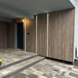 Damiano - Nowoczesna elewacja z drewnianych paneli pionowych z wbudowanym oświetleniem LED, szare płytki chodnikowe i schody, czarne drzwi wejściowe.