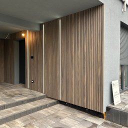 Damiano - Nowoczesna elewacja budynku z drewnianymi panelami pionowymi i wbudowanym oświetleniem LED, widoczne schody wejściowe z kostki brukowej i fragment okna z roletą zewnętrzną.