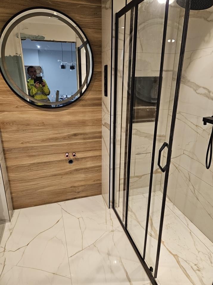 Nowoczesna łazienka z kabiną prysznicową z czarnymi ramami i okrągłym lustrem, odbijającym osobę robiącą zdjęcie, ściany wyłożone płytkami imitującymi drewno i marmur.