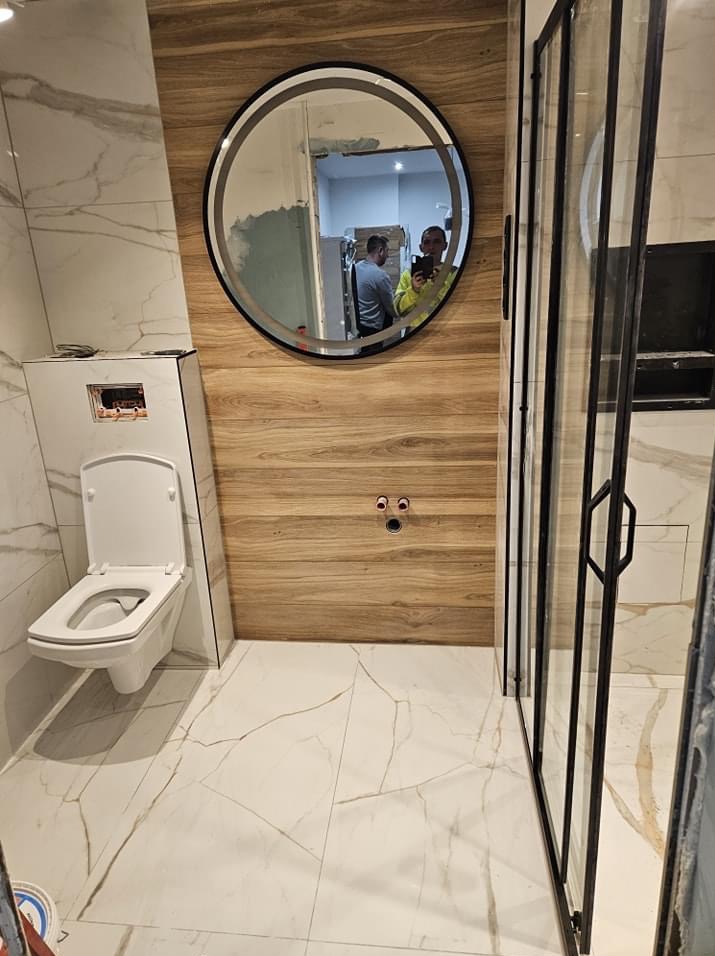 Nowoczesna łazienka w trakcie wykańczania: białe marmurowe płytki podłogowe i ścienne, drewnopodobna okładzina ściany z okrągłym lustrem, podwieszana toaleta i czarna kabina prysznicowa z widocznym...