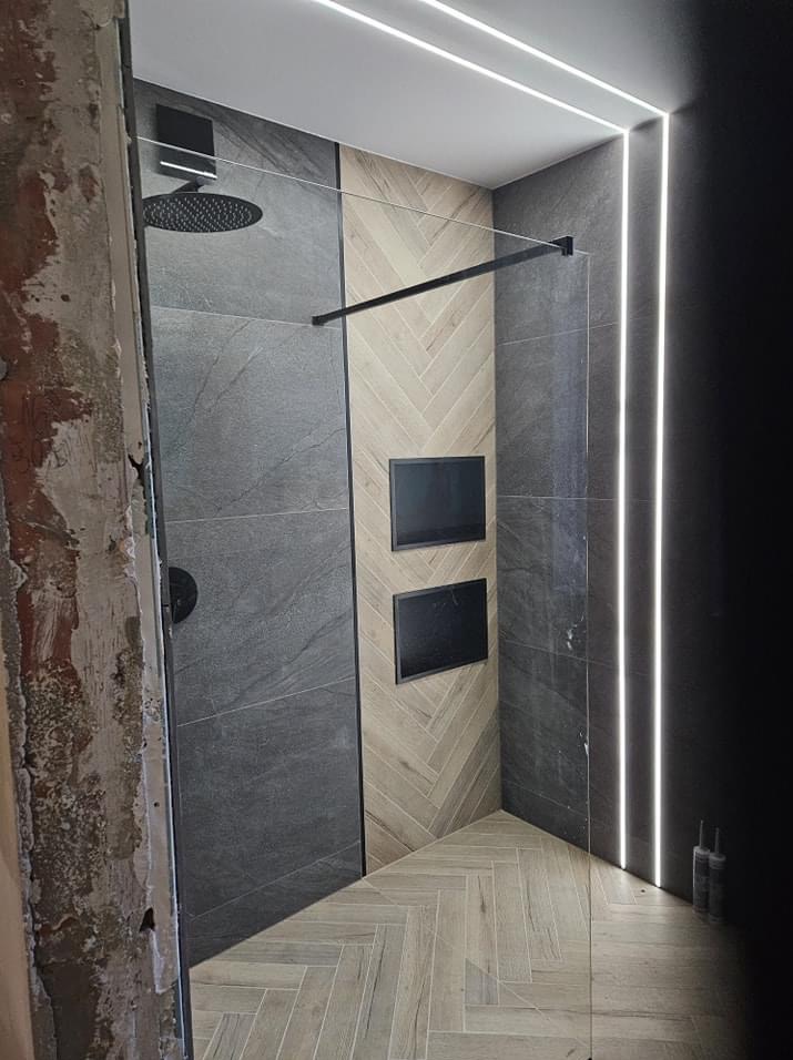 Nowoczesna łazienka w trakcie prac wykończeniowych, widoczne surowe ściany obok kabiny prysznicowej z deszczownicą, podświetlane panele LED i drewnopodobne płytki ułożone w jodełkę.