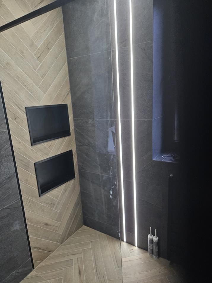 Nowoczesna łazienka z szaro-beżowymi płytkami ułożonymi w jodełkę, podświetlane pionowe pasy LED na ścianie i wnęki w ścianie obłożonej płytkami imitującymi drewno.