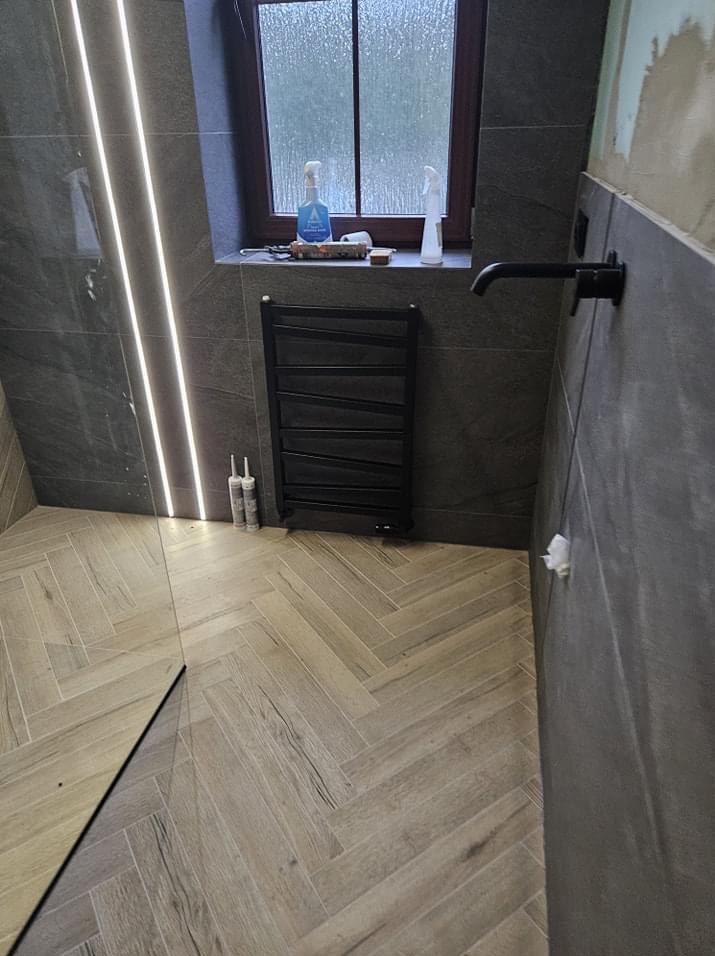 Nowoczesna łazienka w trakcie remontu: szara płytka ścienna, drewnopodobna podłoga ułożona w jodełkę, czarny grzejnik drabinkowy, czarna bateria umywalkowa, listwa LED, silikon sanitarny, okno...
