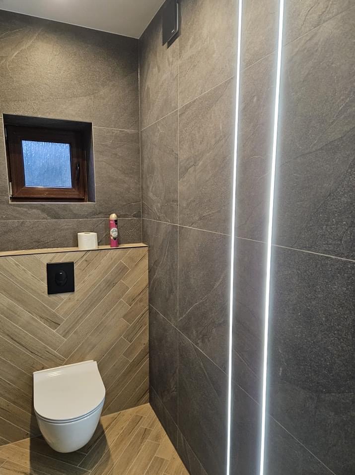 Nowoczesna łazienka z szarymi płytkami imitującymi kamień, podświetleniem LED w pionowych liniach, toaletą podwieszaną i drewnianą obudową stelaża z wzorem jodełki oraz małym oknem.