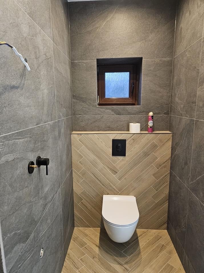 Wykończona łazienka w Jaworznie z szarymi płytkami imitującymi kamień na ścianach, drewnianą obudową stelaża WC ułożoną w jodełkę, małym oknem i czarną armaturą.