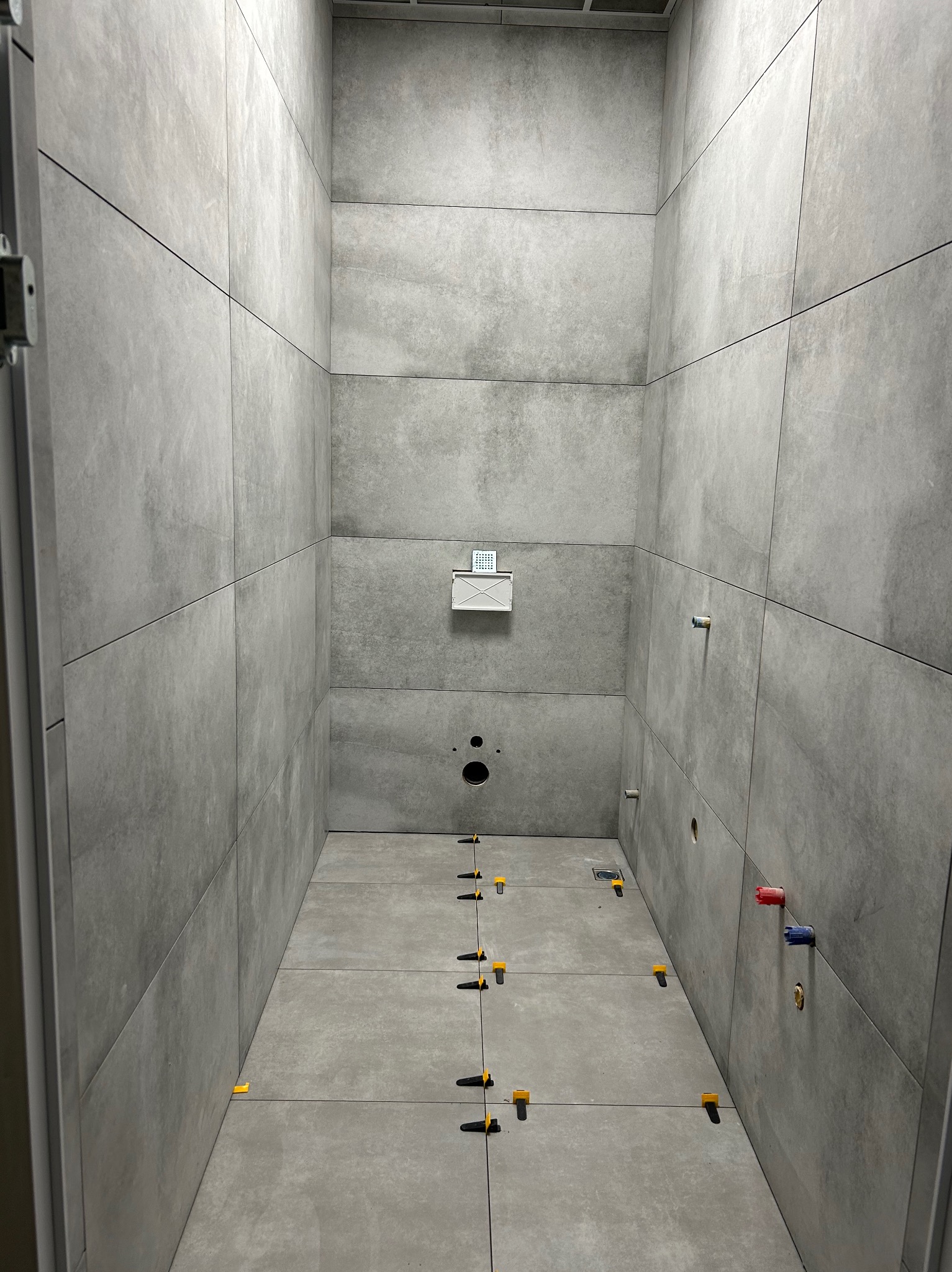 Wnętrze łazienki w trakcie wykańczania, z szarymi płytkami wielkoformatowymi na ścianach i podłodze, widoczne żółte krzyżyki dystansowe, przygotowane punkty instalacyjne wodno-kanalizacyjne.