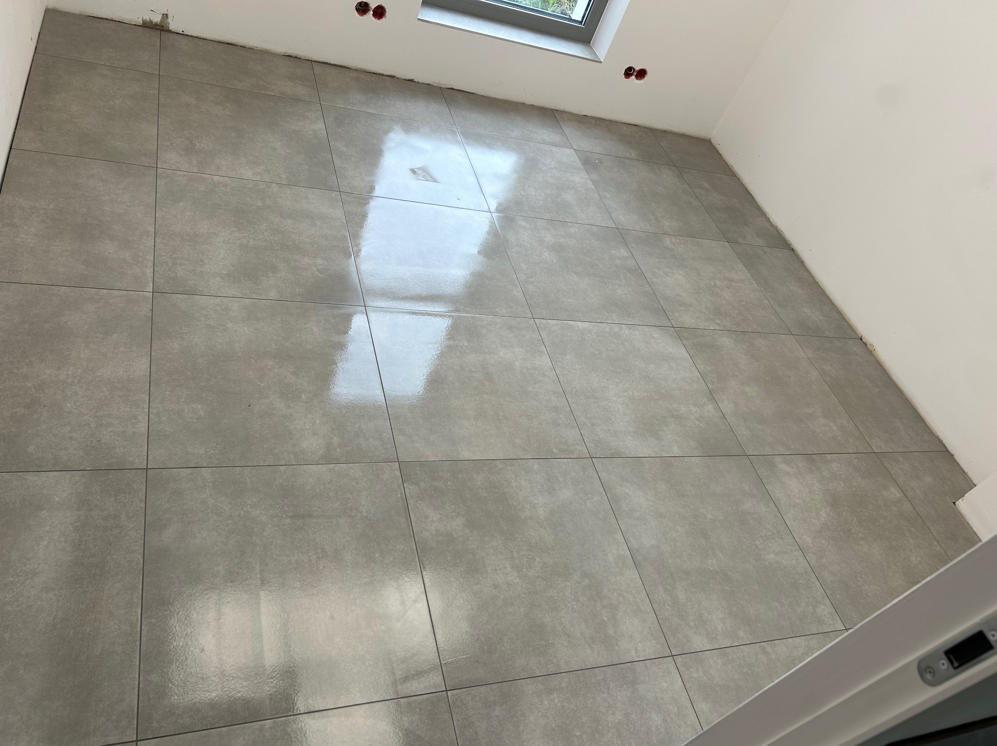 Ułożone szare płytki podłogowe w nowym pomieszczeniu, widoczne gniazdka elektryczne w ścianie i okno w górnej części kadru.