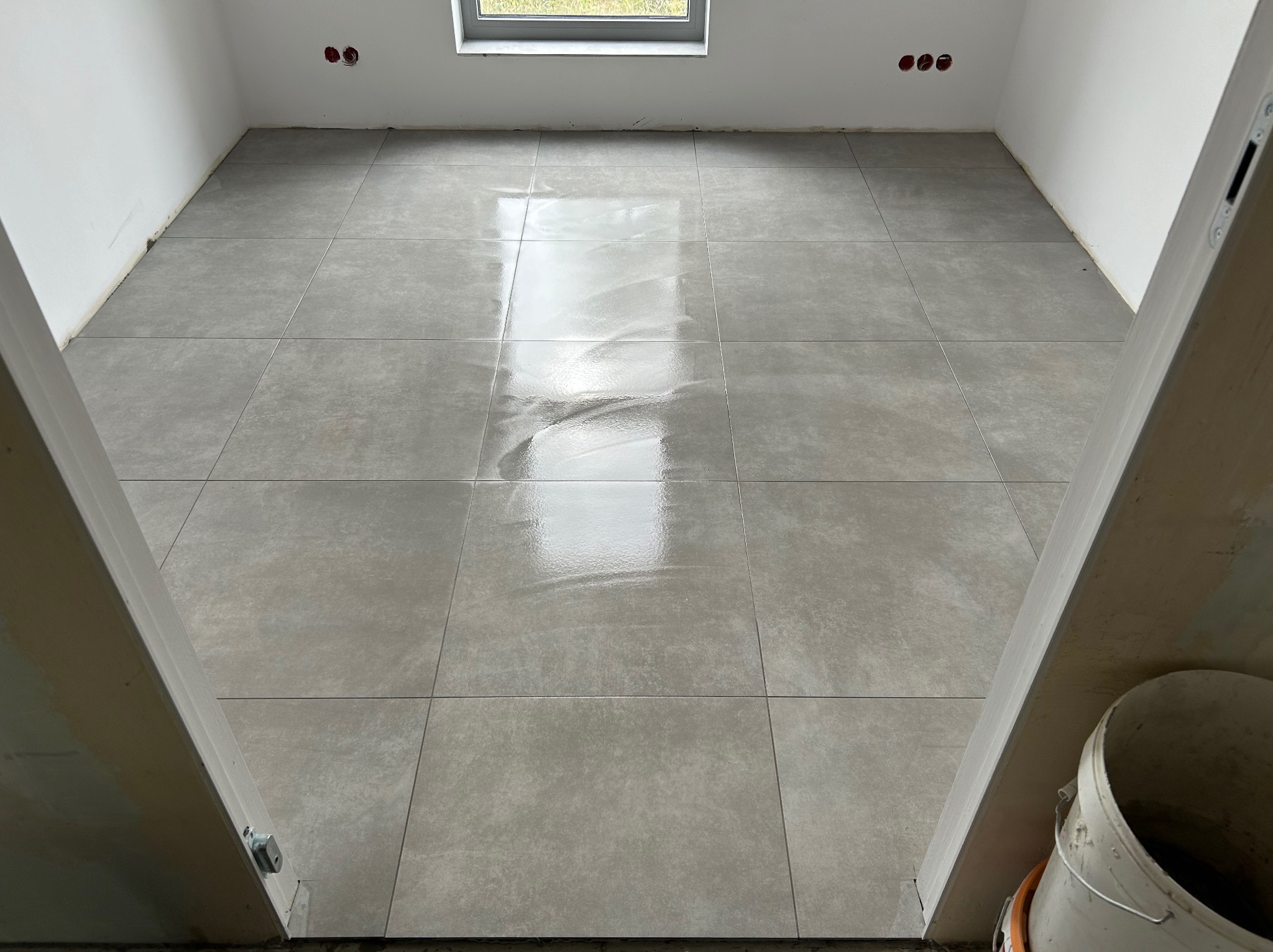 Świeżo ułożone, błyszczące, szare płytki podłogowe w pustym pomieszczeniu, widoczne przez framugę drzwi, z wiadrem przy ścianie.