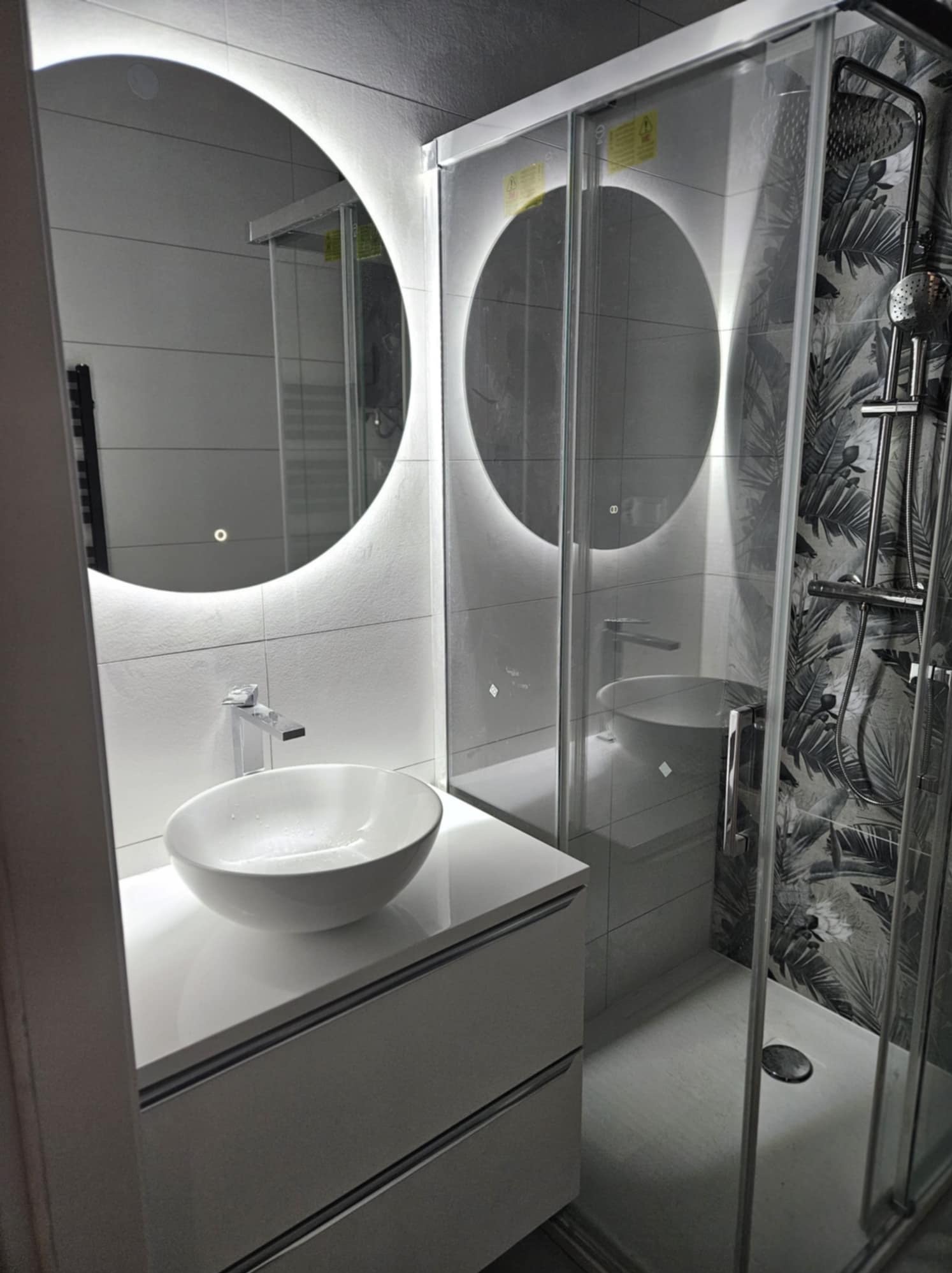 Nowoczesna łazienka z okrągłym lustrem podświetlanym LED, białą umywalką nablatową i kabiną prysznicową z wzorem liści na ścianie.